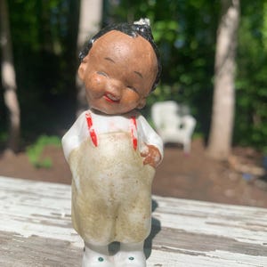 May include: A vintage ceramic figurine of a smiling child with dark skin, black hair, and red suspenders. The child wears a white shirt and tan overalls, and white shoes. The figurine is standing on a weathered wooden surface.