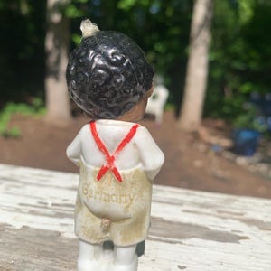 May include: A vintage porcelain figurine of a young boy with black hair, wearing a white shirt with red suspenders and tan overalls. The word "Germany" is printed on the back of the overalls. The figurine is standing on a weathered wooden surface.