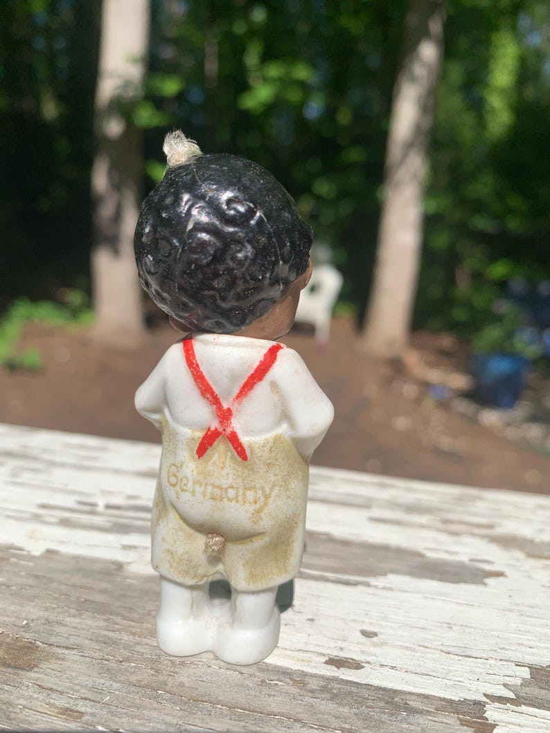 May include: A vintage porcelain figurine of a young boy with black hair, wearing a white shirt with red suspenders and tan overalls. The word "Germany" is printed on the back of the overalls. The figurine is standing on a weathered wooden surface.