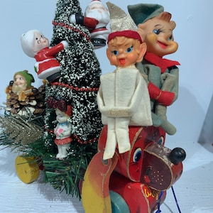 May include: A vintage wooden pull toy shaped like a dog with a red, yellow, and blue paint scheme. The dog is pulling a Christmas tree with a variety of vintage Christmas figurines, including Santa, elves, and a woman in a red dress.