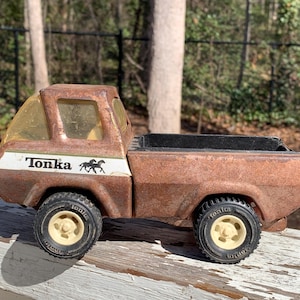 May include: A rusty brown Tonka toy truck with a black bed and white cab. The cab has a clear plastic window and the side of the truck says "Tonka" with a black silhouette of two horses running.