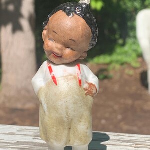 May include: A vintage porcelain figurine of a smiling child with black hair and a white shirt. The child wears tan overalls with red suspenders and white shoes. The figurine has a cheerful expression and is standing outdoors.