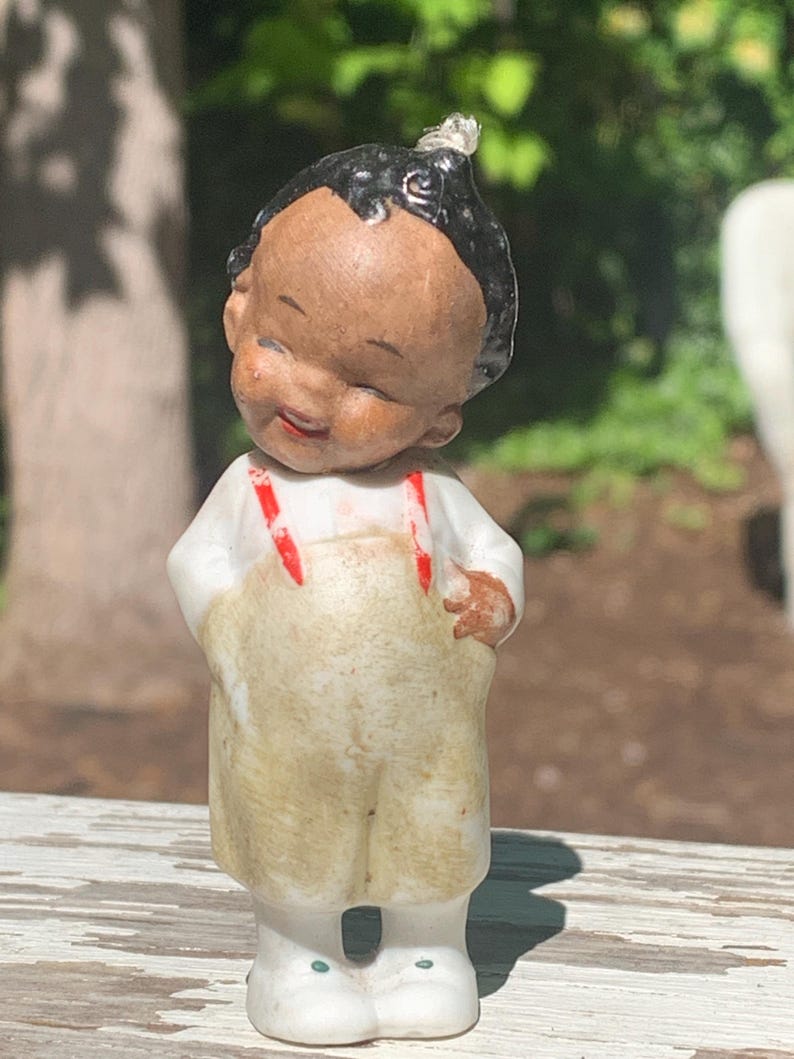 May include: A vintage porcelain figurine of a smiling child with black hair and a white shirt. The child wears tan overalls with red suspenders and white shoes. The figurine has a cheerful expression and is standing outdoors.