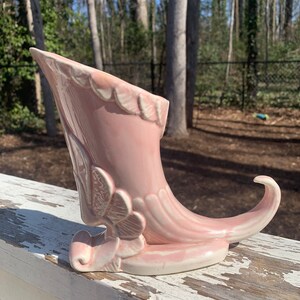May include: A pink ceramic cornucopia vase with a white glaze and floral details. The vase is shaped like a horn with a curled base.
