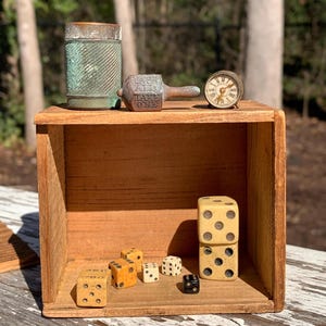 May include: A wooden box contains vintage items. A copper-toned metal container and a small clock sit on top. Inside are several dice, including a pair stacked together. The text "TAKE ONE" is visible on a small metal object.