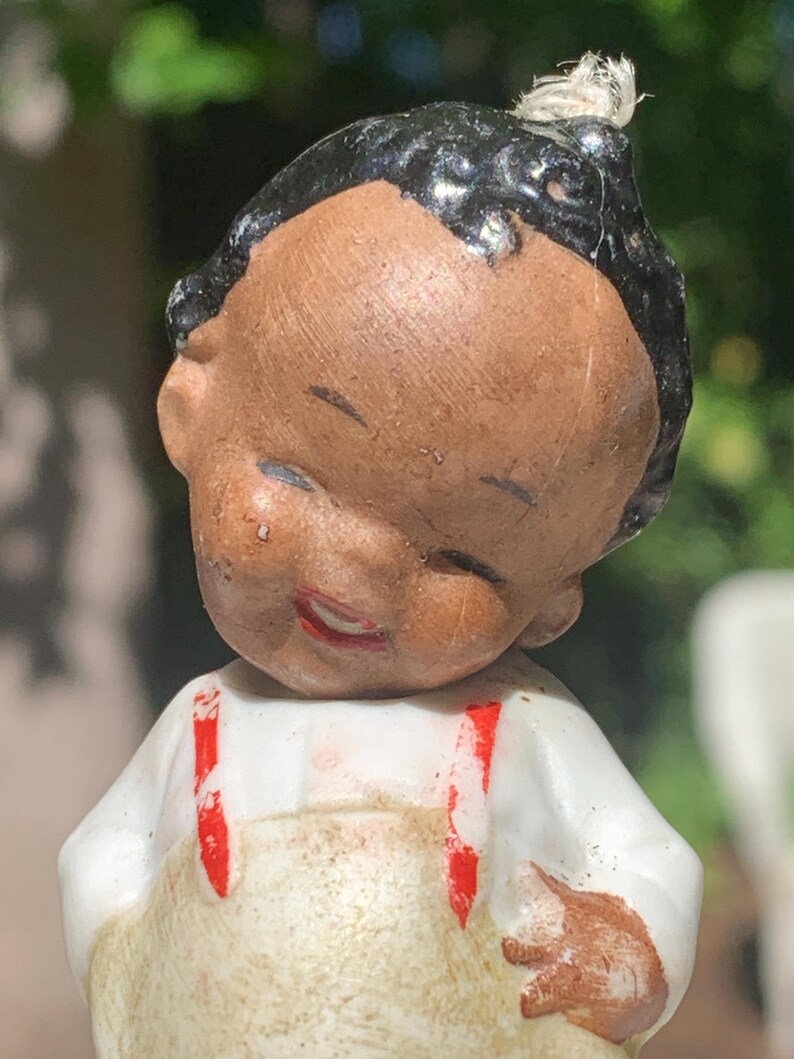 May include: A vintage bisque doll with a smiling face, black hair, and a white shirt with red suspenders. The doll has a tan overall and a small tuft of hair on top of its head. The doll's eyes are closed, and its lips are painted red.