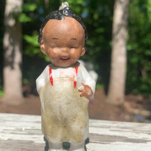 May include: A vintage ceramic figurine of a smiling child with black hair and a brown complexion. The child wears a white shirt, tan overalls with red suspenders, and white shoes. The figurine is standing on a weathered wooden surface.