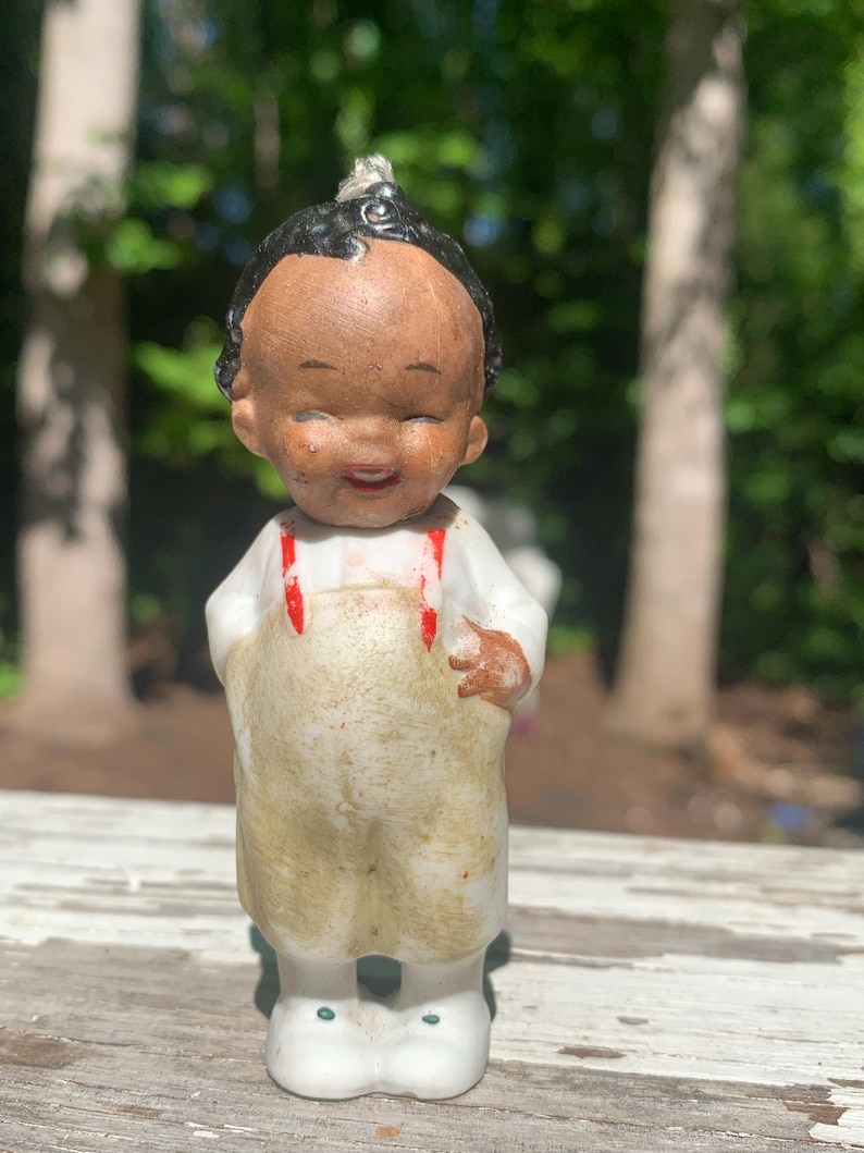 May include: A vintage ceramic figurine of a smiling child with black hair and a brown complexion. The child wears a white shirt, tan overalls with red suspenders, and white shoes. The figurine is standing on a weathered wooden surface.