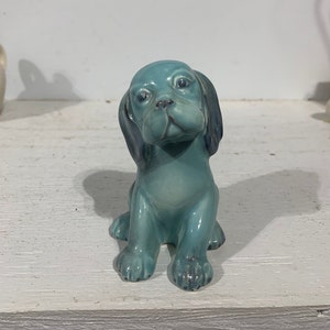 May include: A blue ceramic figurine of a sitting puppy. The puppy has a light blue glaze and dark blue accents around the eyes and ears.