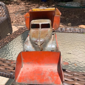 May include: Vintage orange and gray metal toy dump truck. The truck has a weathered appearance with visible rust. The dump bed is orange, and the cab is gray. The truck is sitting on a glass table.