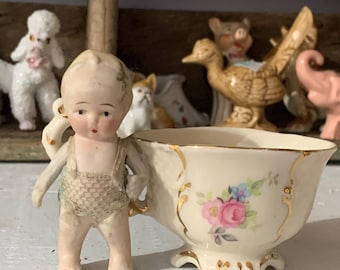 May include: A porcelain doll wearing a swimsuit holds a small teacup with floral decorations. The teacup has a gold rim and a floral pattern.
