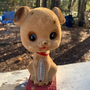 May include: A plush brown dog figurine with large eyes and a red collar sits on a red book. The dog is holding a thermometer with a Celsius and Fahrenheit scale. The thermometer reads 20 degrees Celsius and 68 degrees Fahrenheit.