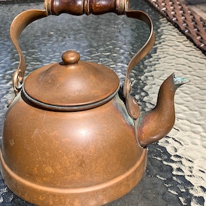 May include: A vintage copper kettle with a wooden handle and a bird-shaped spout. The kettle has a round body, a lid with a small knob, and a curved handle. The copper has a warm, aged patina.