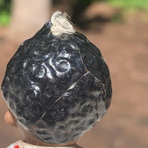 May include: Close-up of a vintage porcelain doll's head, featuring a glossy black hairpiece with a small, white tuft at the top. The doll's head is cracked, revealing its age. The doll is wearing a white shirt with red accents.