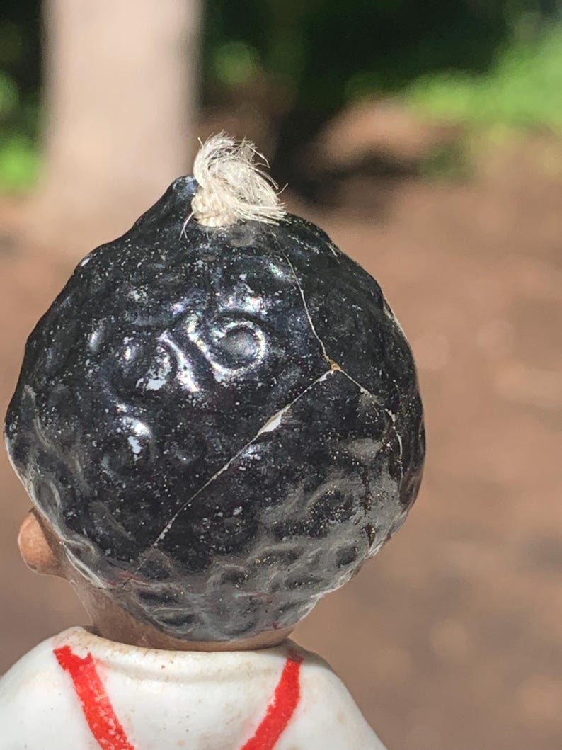 May include: Close-up of a vintage porcelain doll's head, featuring a glossy black hairpiece with a small, white tuft at the top. The doll's head is cracked, revealing its age. The doll is wearing a white shirt with red accents.