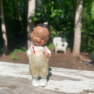 May include: A vintage ceramic figurine of a child with dark skin, wearing tan overalls with red suspenders and white shoes. The figurine has a smiling expression and a small topknot. The background is a blurred outdoor scene.