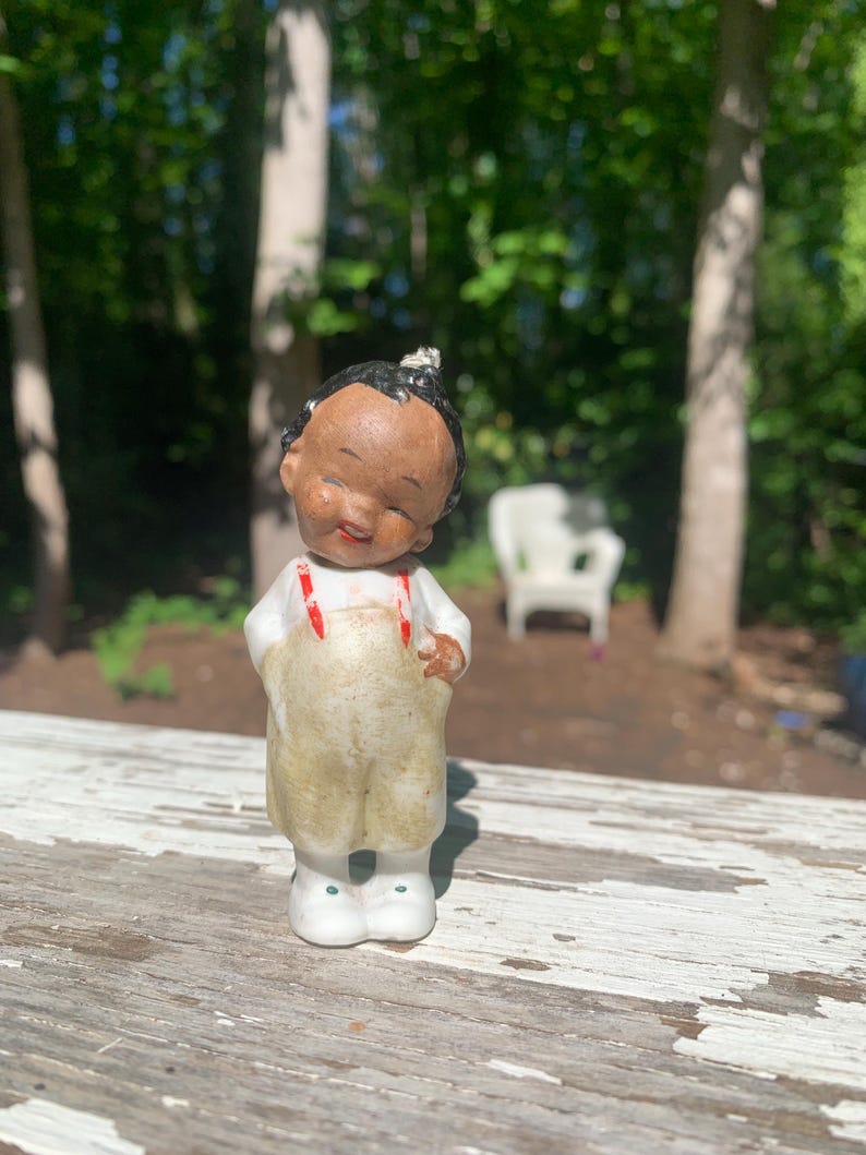 May include: A vintage ceramic figurine of a child with dark skin, wearing tan overalls with red suspenders and white shoes. The figurine has a smiling expression and a small topknot. The background is a blurred outdoor scene.