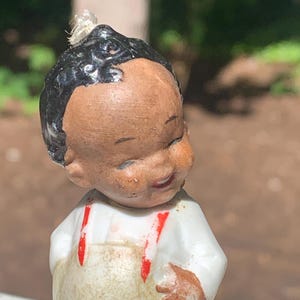 May include: A vintage figurine of a smiling child with dark hair and brown skin. The figurine wears a white shirt with red suspenders and a tan overall. The figurine's hand is resting on the overall.