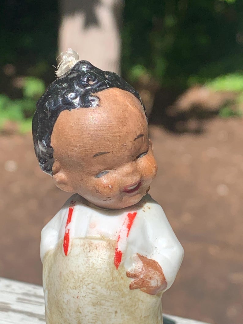 May include: A vintage figurine of a smiling child with dark hair and brown skin. The figurine wears a white shirt with red suspenders and a tan overall. The figurine's hand is resting on the overall.