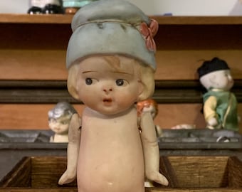 May include: A porcelain doll with a blue hat and pink flower accents. The doll has a light pink complexion and is wearing a white dress. The doll is missing its arms and is posed with its hands on its hips.