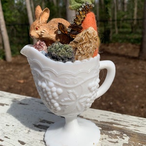 May include: A white milk glass pitcher with a grape design, filled with a small brown rabbit, a carrot, and other decorative items.