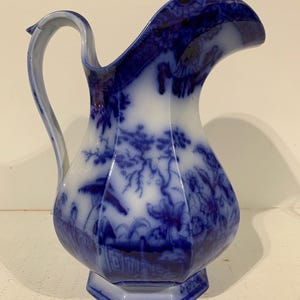 May include: A white ceramic pitcher featuring a deep blue transferware pattern of floral and botanical motifs. The vessel has a faceted, hexagonal body, a curved handle, and a wide pouring spout, with the blue design wrapping around the exterior.