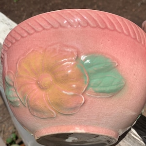 May include: A pink ceramic bowl with a raised floral design. The flower is peach and yellow, with green leaves. The rim has a rope-like texture. The bowl has a crackled finish.