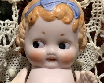 May include: A vintage porcelain doll head with a blue ribbon. The doll has blonde hair, rosy cheeks, and black eyes. The doll's arms are visible. The doll is set against a background of lace and patterned fabric.