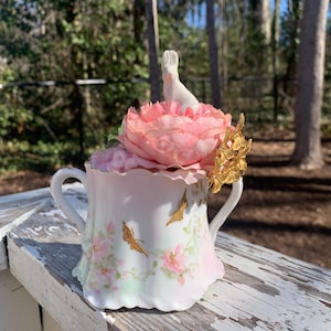 May include: A white porcelain teacup with a floral design and gold accents. The cup is decorated with pink flowers and butterflies. A white porcelain rabbit figurine sits on top of a pink flower.