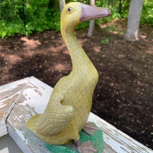 May include: A yellow ceramic goose figurine with a brown beak and green grass base. The goose has detailed feathers and a realistic appearance.