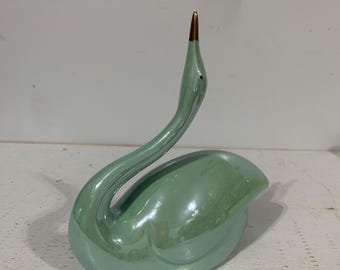 May include: A glossy, light mint green ceramic swan figurine with a smooth, iridescent finish. The swan features an elongated, curved neck and a pointed beak tipped with a metallic gold accent, capturing a graceful posture in a stationary position.