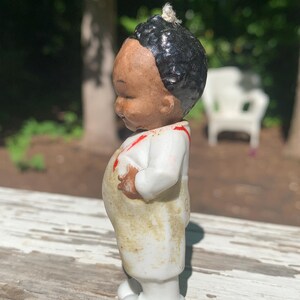 May include: A vintage porcelain figurine of a young child. The figurine has a brown face, black hair, and is wearing a white outfit with red accents. The figurine is standing on a white base.