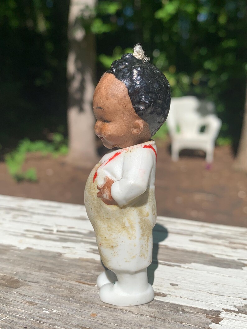 May include: A vintage porcelain figurine of a young child. The figurine has a brown face, black hair, and is wearing a white outfit with red accents. The figurine is standing on a white base.