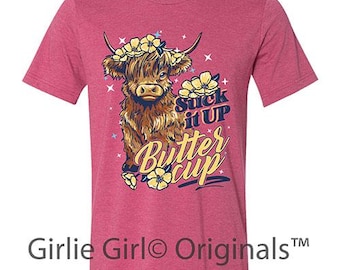 May include: A short-sleeved, heathered pink t-shirt featuring a Highland cow illustration with a floral crown and the text "Suck it Up Buttercup." The shirt also has the text "Girlie Girl Originals™" at the bottom.
