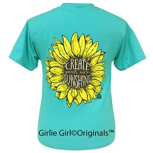 May include: A turquoise blue t-shirt with a yellow sunflower graphic and the text "Create your own sunshine" in the center. The shirt is made by Girlie Girl Originals.