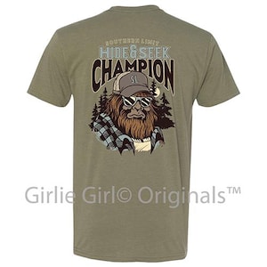 May include: Olive green t-shirt with a graphic of a Bigfoot wearing sunglasses and a hat. The text on the shirt reads "SOUTHERN LIMIT HIDE & SEEK CHAMPION". The brand name "Girlie Girl Originals" is printed at the bottom.