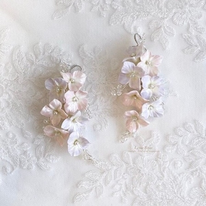 Peut inclure: Une paire de boucles d'oreilles délicates avec des grappes de fleurs d'hortensias rose pâle et blanches. Les fleurs sont en pâte polymère et ont une finition nacrée. Les boucles d'oreilles sont suspendues à des crochets argentés.