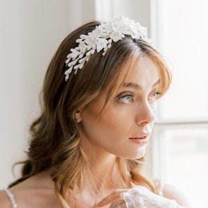 May include: A white floral headband with delicate white flowers and leaves. The headband is worn by a woman with long, wavy brown hair.