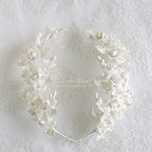 May include: White floral bridal hair crown with delicate white flowers and leaves. The crown is made of a flexible wire and is designed to fit comfortably on the head. The crown is adorned with the text "Lena Rom Couture Bridal Accessories".