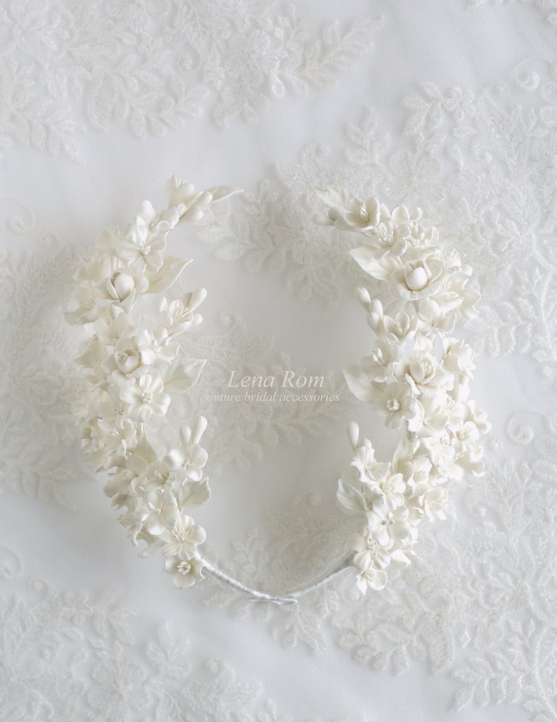 May include: White floral bridal hair crown with delicate white flowers and leaves. The crown is made of a flexible wire and is designed to fit comfortably on the head. The crown is adorned with the text "Lena Rom Couture Bridal Accessories".