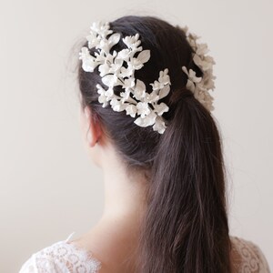May include: White floral hair comb with delicate white flowers and leaves. The comb is worn in a woman's hair, creating a romantic and elegant look.