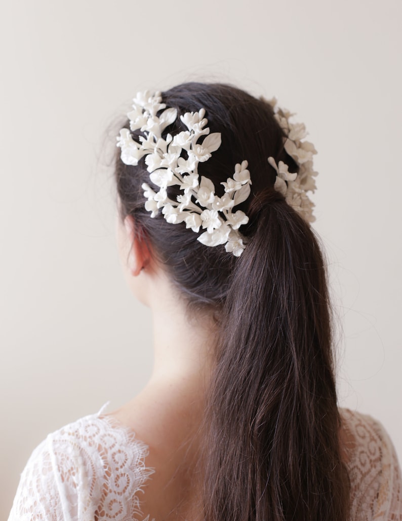 May include: White floral hair comb with delicate white flowers and leaves. The comb is worn in a woman's hair, creating a romantic and elegant look.
