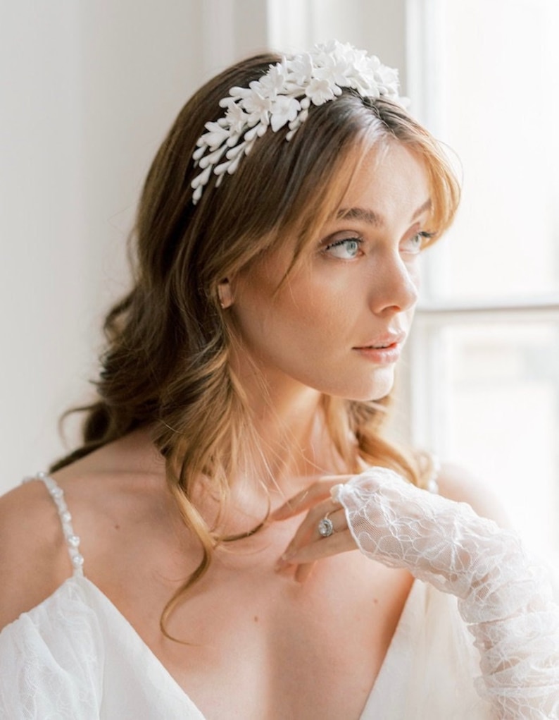 May include: A white floral headband with delicate white flowers and leaves. The headband is worn by a woman with long, wavy brown hair.