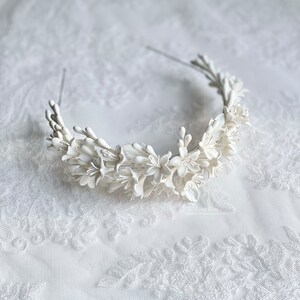 May include: A white floral headband with delicate white flowers and leaves. The headband is made of a thin wire and is designed to be worn around the head.