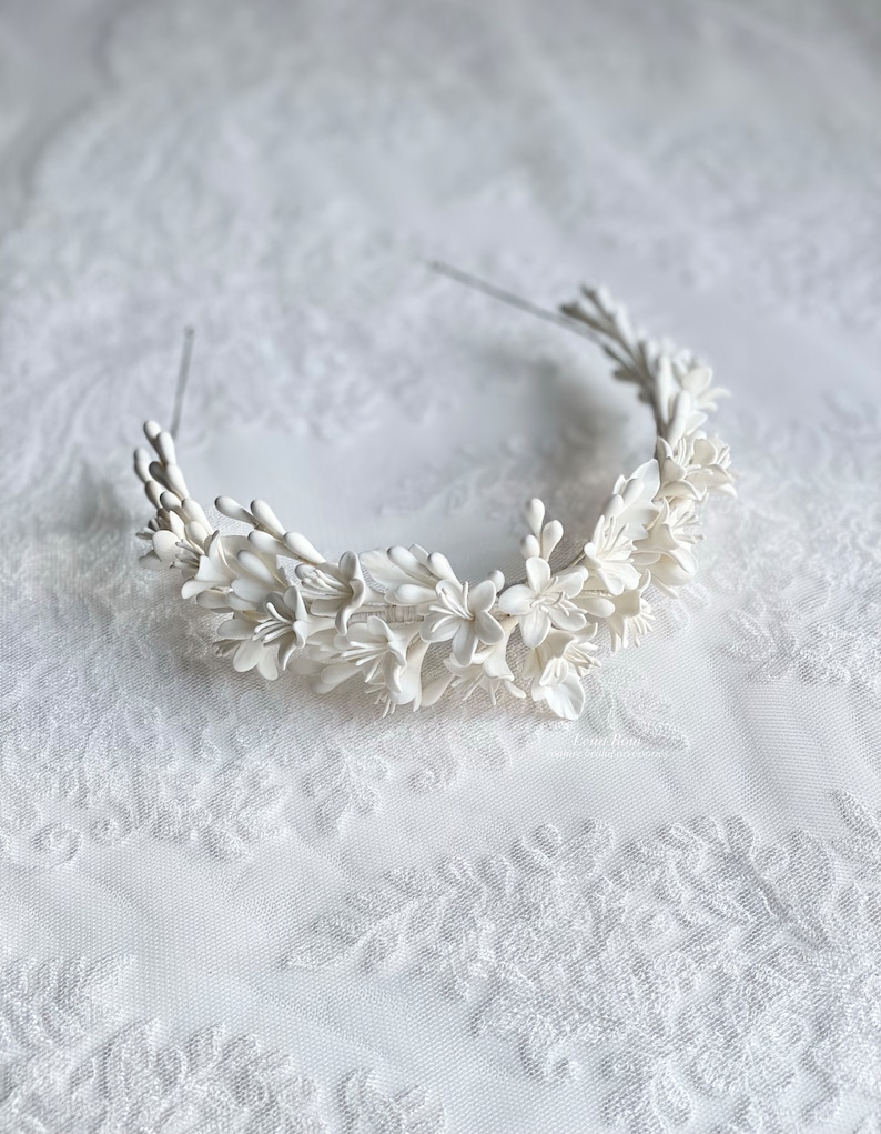 May include: A white floral headband with delicate white flowers and leaves. The headband is made of a thin wire and is designed to be worn around the head.