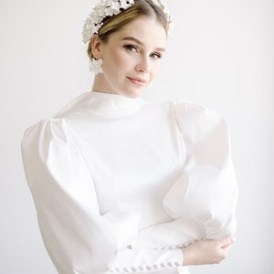 May include: A woman wearing a white dress with a high neckline and puffy sleeves. She is wearing a white flower crown and white flower earrings.