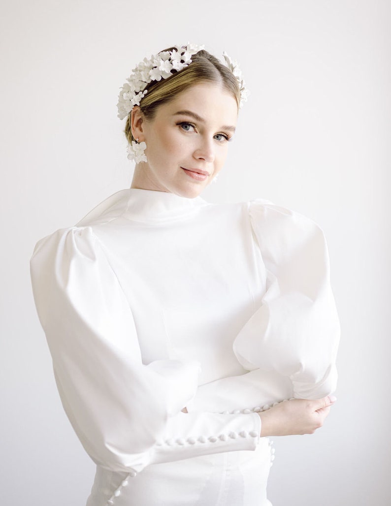 May include: A woman wearing a white dress with a high neckline and puffy sleeves. She is wearing a white flower crown and white flower earrings.