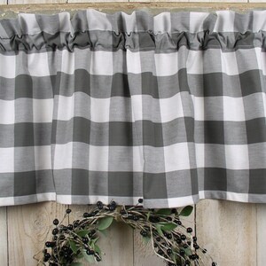 May include: A gray and white buffalo check valance with a ruffled top edge. The curtain is hanging in front of a wooden background. A wreath with black berries and green leaves is below the curtain.