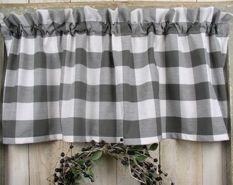May include: A gray and white buffalo check valance with a ruffled top edge. The curtain is hanging in front of a wooden background. A wreath with black berries and green leaves is below the curtain.