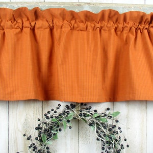 May include: A burnt orange window valance featuring a subtle, thin dark grid pattern. The fabric is gathered at the top with a rod pocket header, creating soft, vertical folds that drape downward across the width of the rectangular textile.